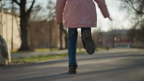 close-up of a young girl's legs in motion, wearing blue jeans and black shoes, running on a sunlit park pathway with blurred trees and a building background