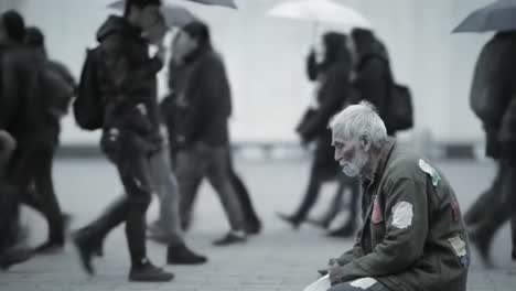 a lonely homeless man sits on the ground in a city square, surrounded by rushing pedestrians who hold umbrellas. rain creates a somber atmosphere while people walk by without noticing him.