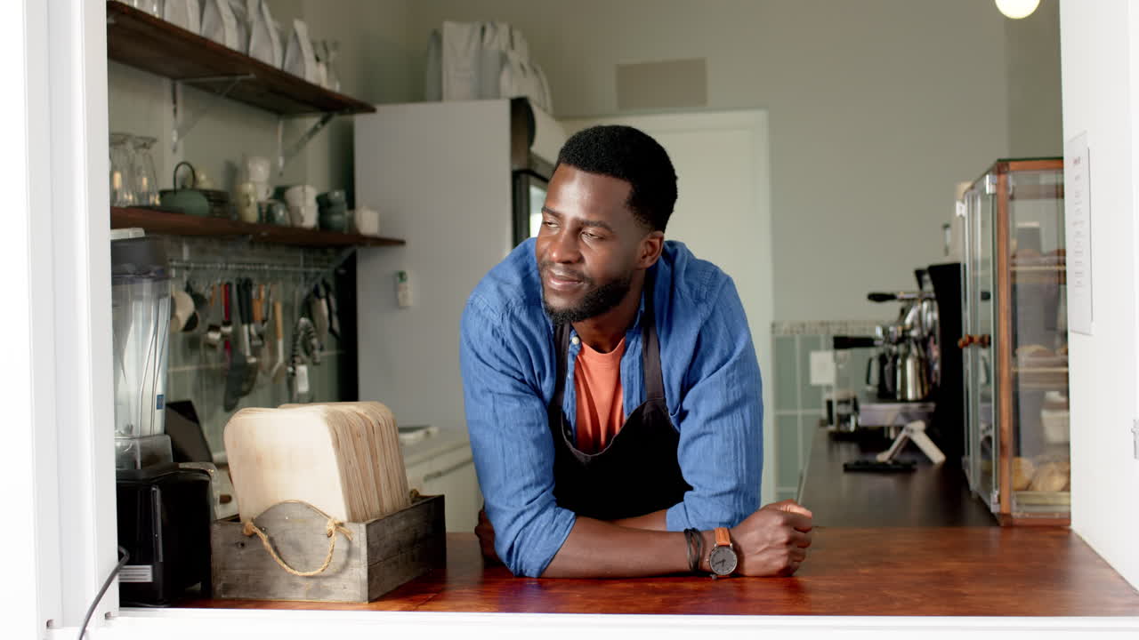 A young African American male barista leans on a cafe counter, smiling at the camera