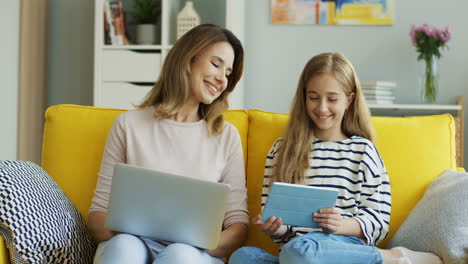 blonde mutter und tochter sitzen auf einer gelben couch, während sie laptop und tablet benutzen und im wohnzimmer reden