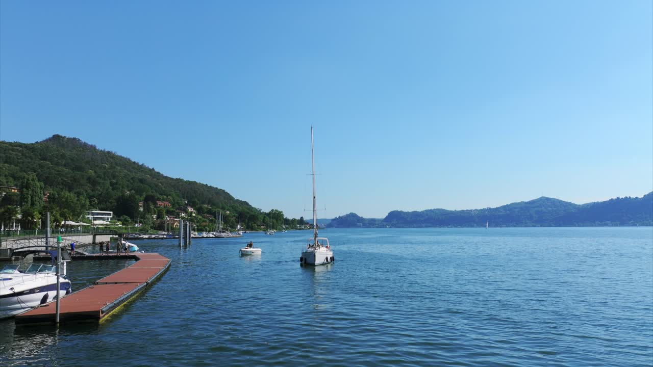 Aerial view showing a white sailboat with sails down, anchored near a wooden dock on a calm lake, surrounded by hills under a clear blue sky on a bright sunny day