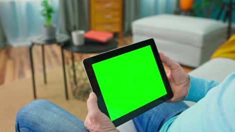 primer plano de una tableta digital con una pantalla de simulacro verde en blanco sostenida por un anciano adulto relajándose en una habitación acogedora en casa. la cámara se aleja de la tableta