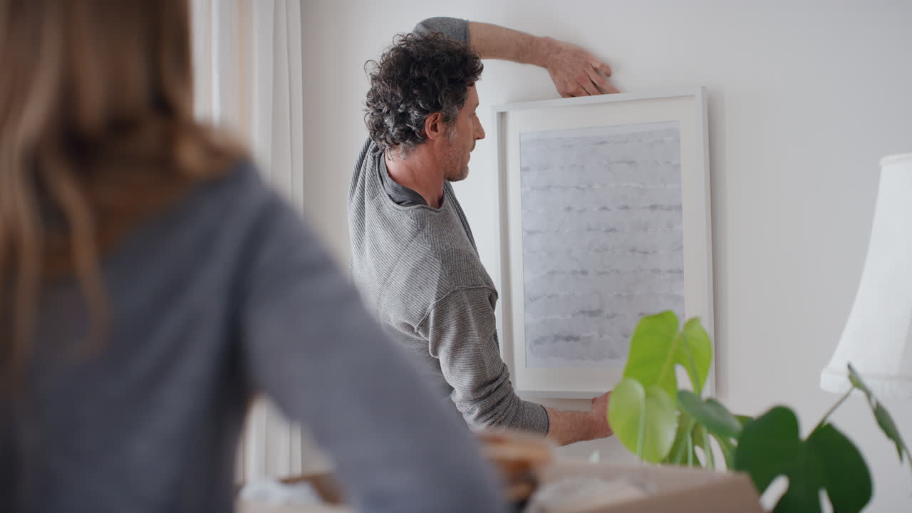 행복한 커플이 새로운 집으로 이사하고 벽에 그림을 걸고 집을 장식하고 팀워크를 즐기고 있습니다.