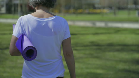 rear view of woman walking in park, holding yoga mat in hand