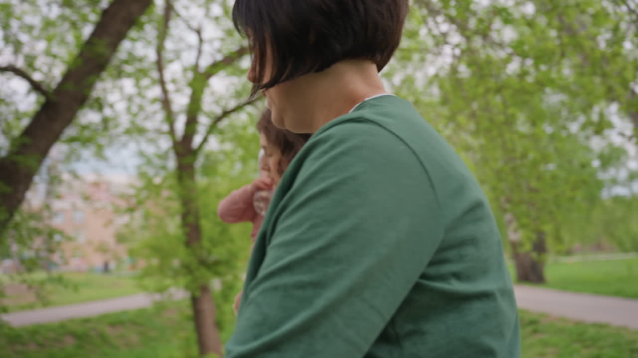 Strolling Women Carry Infant Nearby, Two Females Strolling Through Green Park Carrying Their Child, Pair Of Women Ambling With Their Infant Amid Leafy Neighborhood Surroundings And Tall Buildings