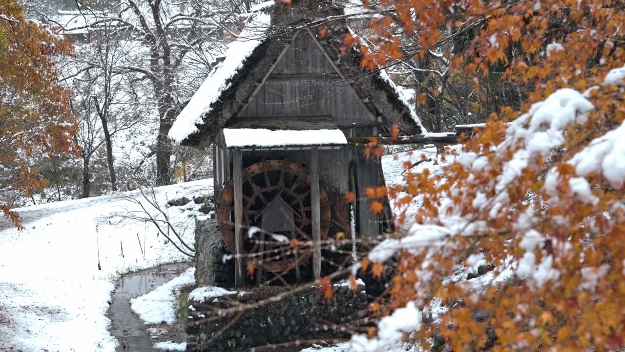 A traditional water mill stands in Shirakawa-go, Japan, surrounded by vibrant red autumn foliage as gentle snowflakes fall, blending two beautiful seasons.