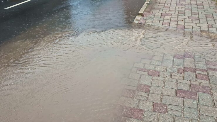 Rainwater overflowing onto a paved sidewalk as runoff from the street creates shallow flooding across the patterned pavement