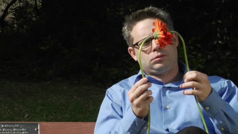 Ein-Mann-Sitzt-Allein-Auf-Einer-Parkbank-Und-Hält-Zwei-Blumen-In-Der-Hand