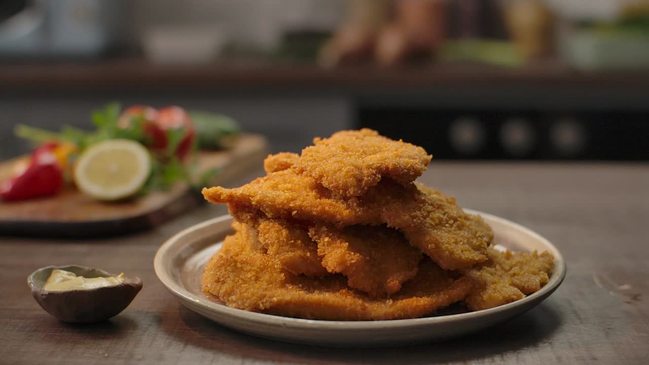 A perfectly fried, golden-brown chicken schnitzel is added to a towering stack of crispy cutlets on a rustic kitchen table