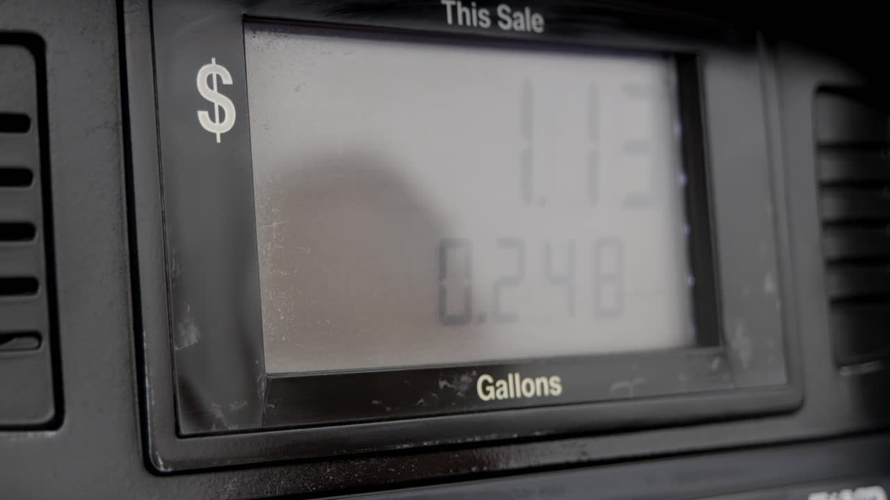 Schermo della pompa di benzina che si sta riempiendo e pompa lentamente fino a meno di un gallone, con il riflesso sullo schermo di una persona con uno chignon.