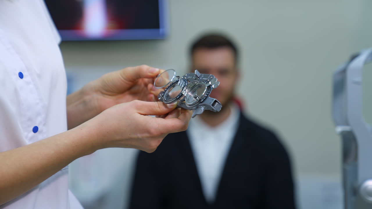 An eye doctor is going to check the eyesight and puts different lenses in special glasses for a young man. Young man on blurred background.