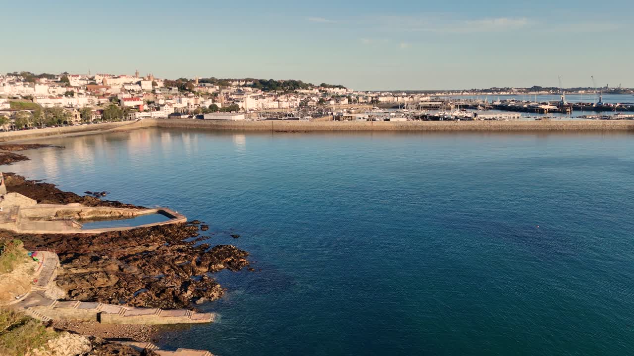 Volo sopra le piscine naturali di acqua salata e la baia di Havelet con l'alta marea, verso il porto di St Peter Port, con mare calmo sotto il sole del tardo pomeriggio, vicino a scogliere boscose e l'isola di Herm in lontananza