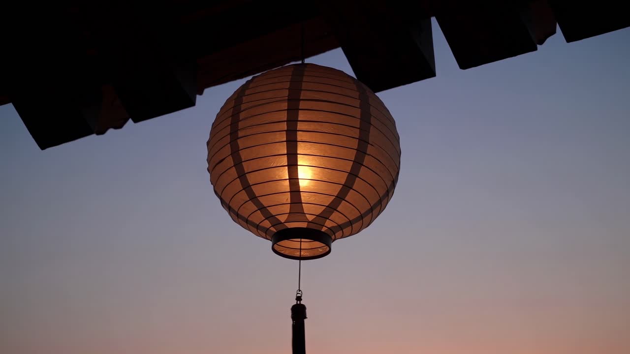 Illuminated round paper lanterns hanging from a wooden roof, casting a warm glow and creating a serene atmosphere during a stunning sunset, evoke tranquility and peace