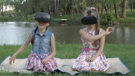 happy children, two little girls, wearing virtual reality glasses, play and draw on a virtual board, sitting on the grass in the park on a summer evening. they smile and laugh. the concept. 4k.