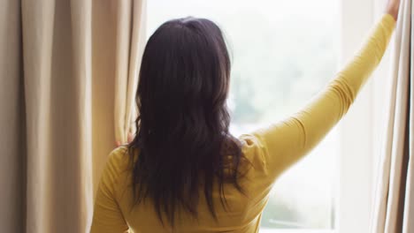 Back-view-of-biracial-woman-revealing-curtains-and-looking-outside-window