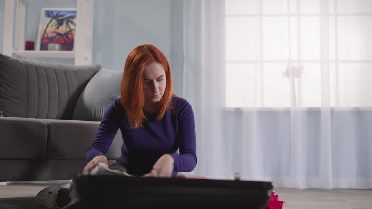 рыжеволосая женщина берет одежду из открытого черного чемодана.