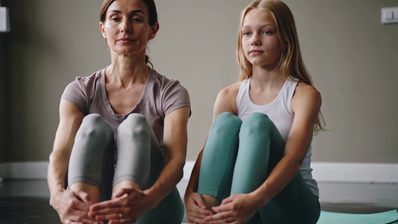 La telecamera si allontana e si sposta verso l'alto, madre e figlia adolescente in abbigliamento sportivo si tengono gli stinchi e respirano su tappetini da yoga in studio.