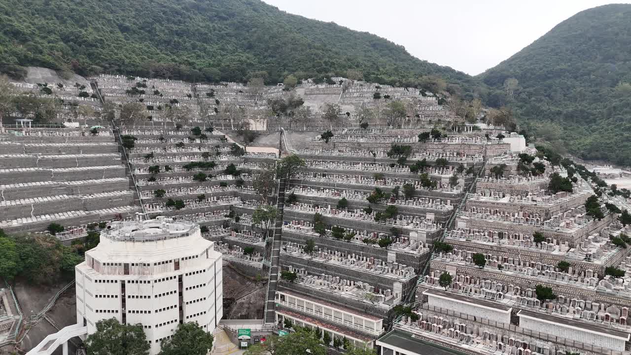 Riprese aeree con drone del Cimitero di Pok Fu Lam a Hong Kong, che mostrano tombe sulla collina, monumenti storici e il paesaggio urbano che si fonde con lo sfondo della città metropolitana.
