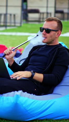 handsome man in black clothes and sunglasses relaxing in bean bag chair smoking hookah. male working on his laptop outdoors in a relaxed atmosphere. vertical video