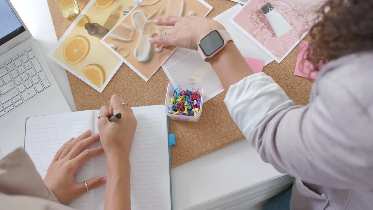 El diseñador de la derecha busca una chincheta y organiza imágenes en un moodboard, mientras el de la izquierda toma notas.