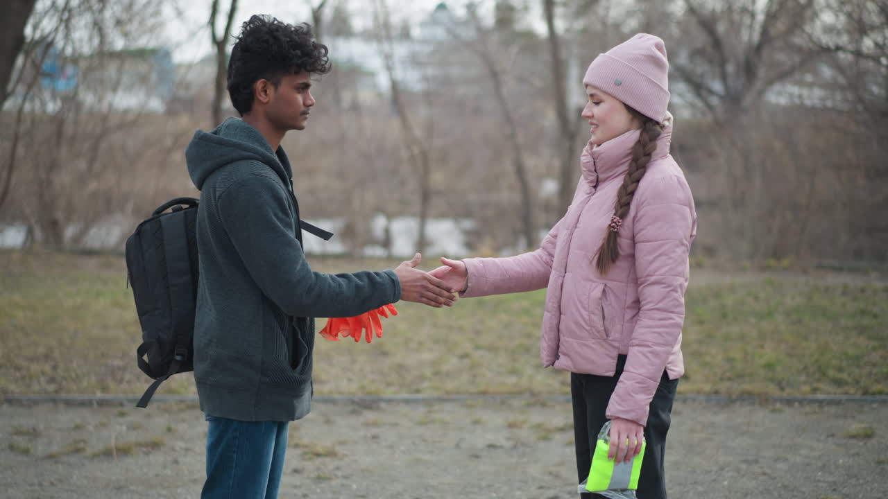 Black guy wearing dark gray hoodie holding orange gloves talking to woman in pink jacket and beanie holding neon safety vest, standing outdoors in park during early spring with bare trees and cloudy sky