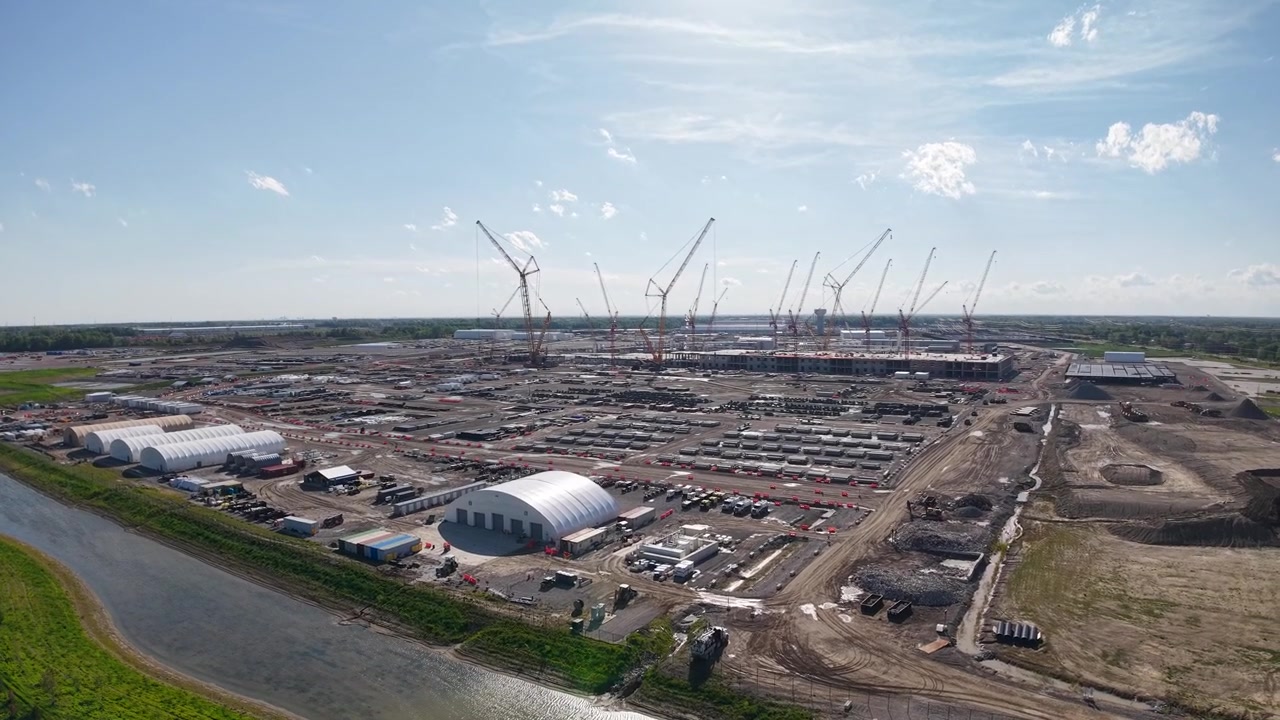 Low aerial view past a retention pond toward cranes covering a vast industrial construction site