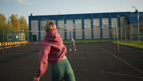 vista trasera de un principiante sirviendo voleibol en una cancha al aire libre con personas borrosas en el fondo, la red de voleibol, el edificio y el campo son visibles mientras el jugador se prepara para el servicio