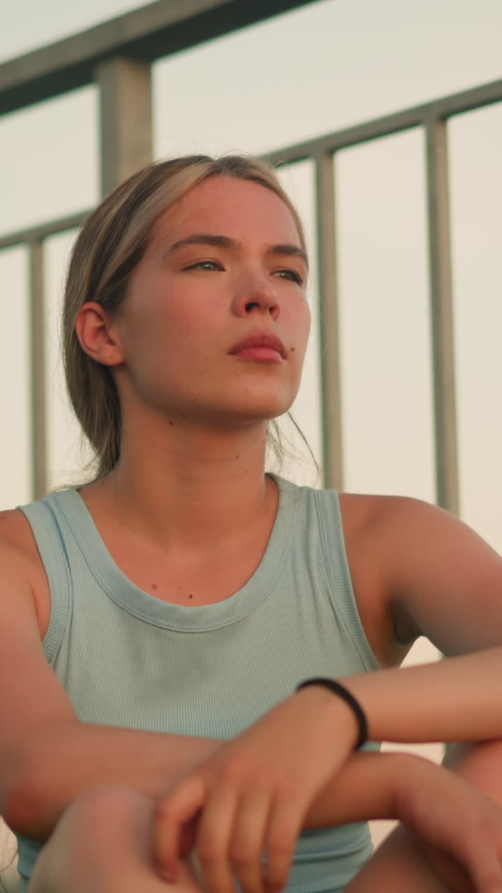joven sentada al aire libre cerca de la barandilla de hierro al anochecer, tocando su cabello con expresión serena, con un brazalete negro en la muñeca, los brazos cruzados sobre las rodillas mientras mira pensativo en la distancia