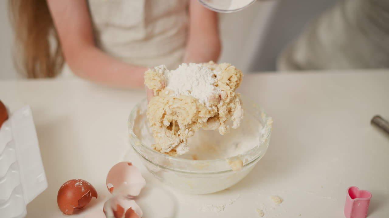 Little kid mixing dough in glass bowl while adult pours flour into hand, assisting with baking process, surrounded by eggshells, flour dust, and baking tools on white table in bright cozy kitchen