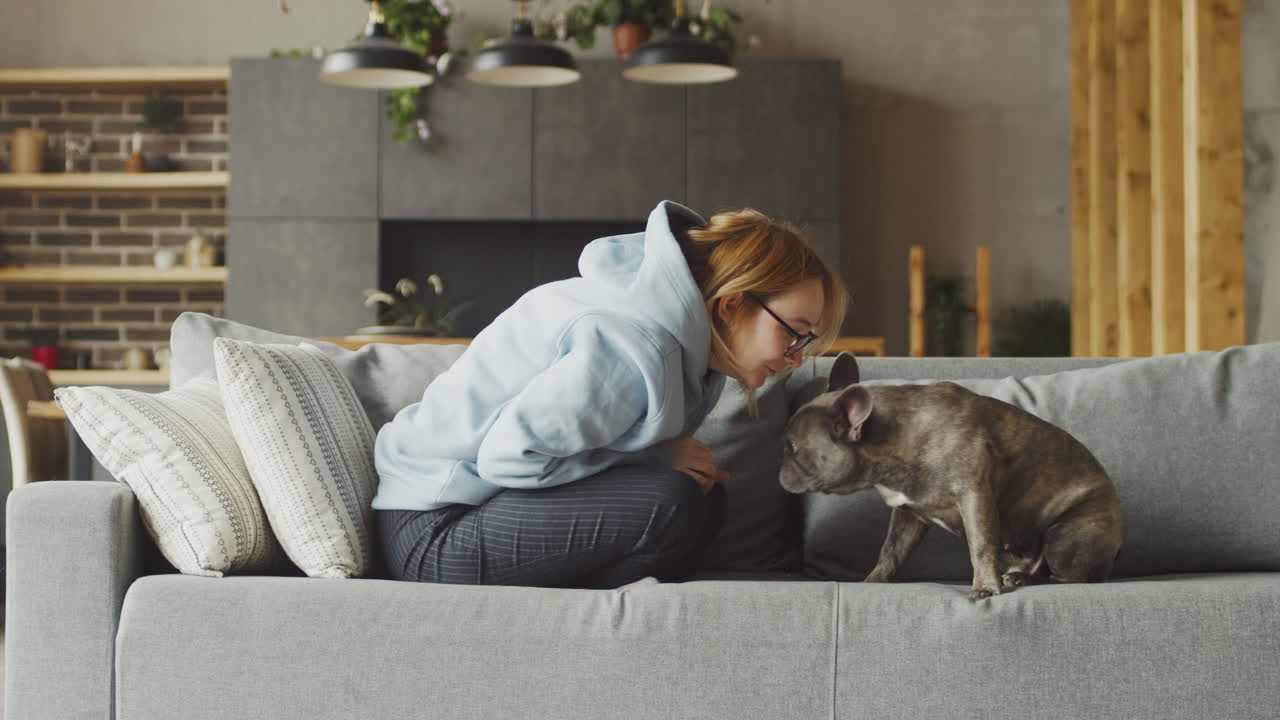 рыжеволосая женщина ласкает свою собаку породы бульдог, когда они сидят на диване в гостиной дома