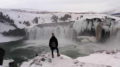 aerial orbit view of tourist standing at the edge of snowy cliff at goðafoss waterfall iceland, revealing spectacular winter landscape around