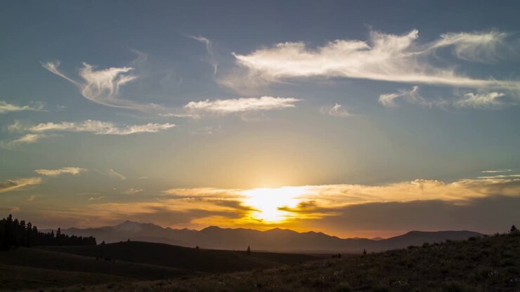 Sole che sorge e cielo luminoso che si alzano sopra il crinale di colline erbose, retroilluminando nuvole cirro.