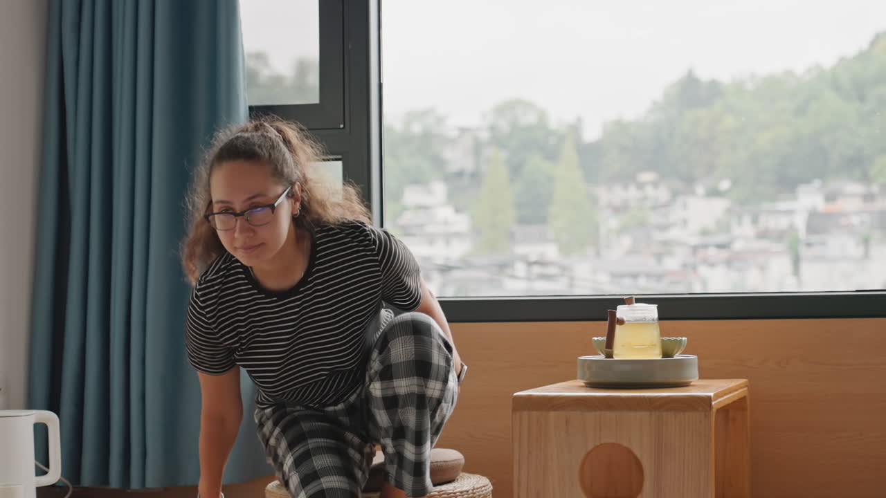 Giovane donna al finestrino dell'hotel che sorseggia il tè, viaggiatrice solitaria che si gode una mattinata tranquilla in una piccola camera d'hotel con vista sulla città, vestiti a quadri, tavolino in legno con vassoio, tazza e teiera in ceramica, tende blu che incorniciano.