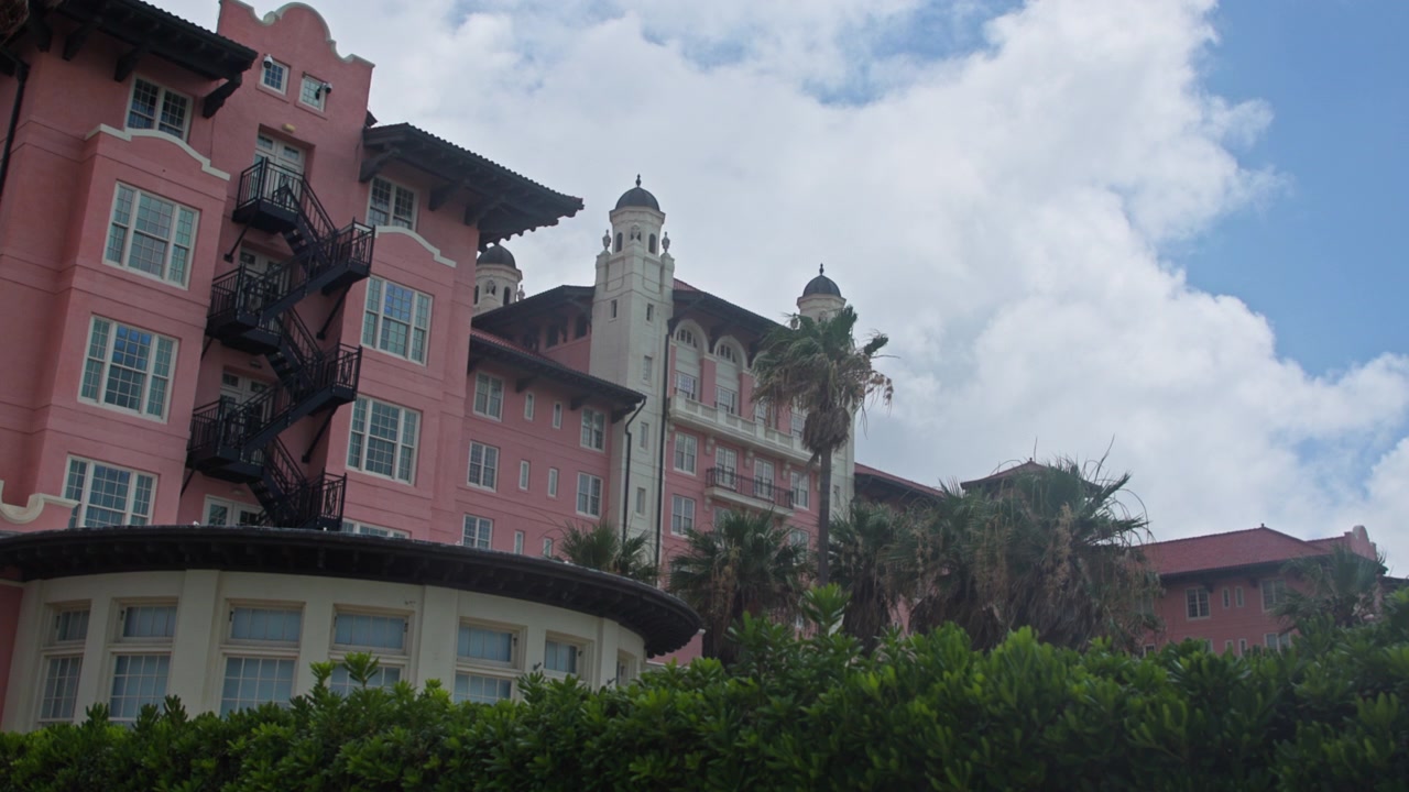 Palme lussureggianti costeggiano un suggestivo hotel storico rosa in Texas, USA. L'ampio angolo cattura l'architettura in stile Revival Mediterraneo, nuvole drammatiche, vegetazione vibrante e un'atmosfera calda diurna.