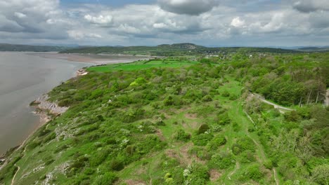 volando sobre el escarpado arbusto verde salpicado de la costa hacia campos de patchwork con horizonte nublado en un día brillante de primavera en el punto de jenny brown, silverdale, lancashire, inglaterra, reino unido