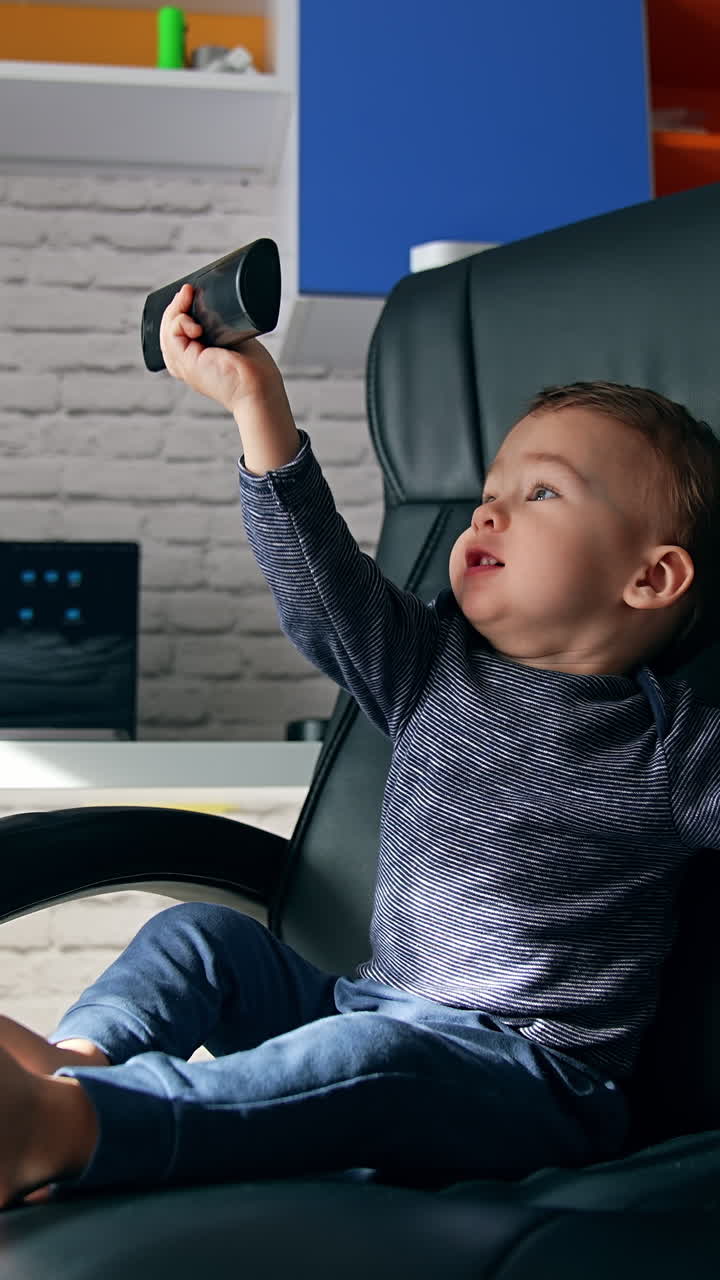 Beautiful baby in the office chair with remote control in hands. Kid turns to the table with laptop and puts the remote control there. Vertical video