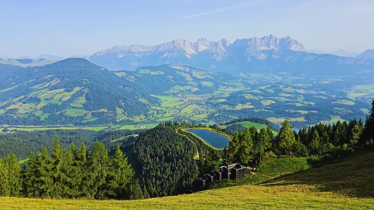 Ampio panorama alpino estivo con prati verdi, pinete, villaggi di valle, un bacino montano e cime aspre sotto un cielo azzurro limpido, ideale per riprese di viaggio europee nelle Alpi.