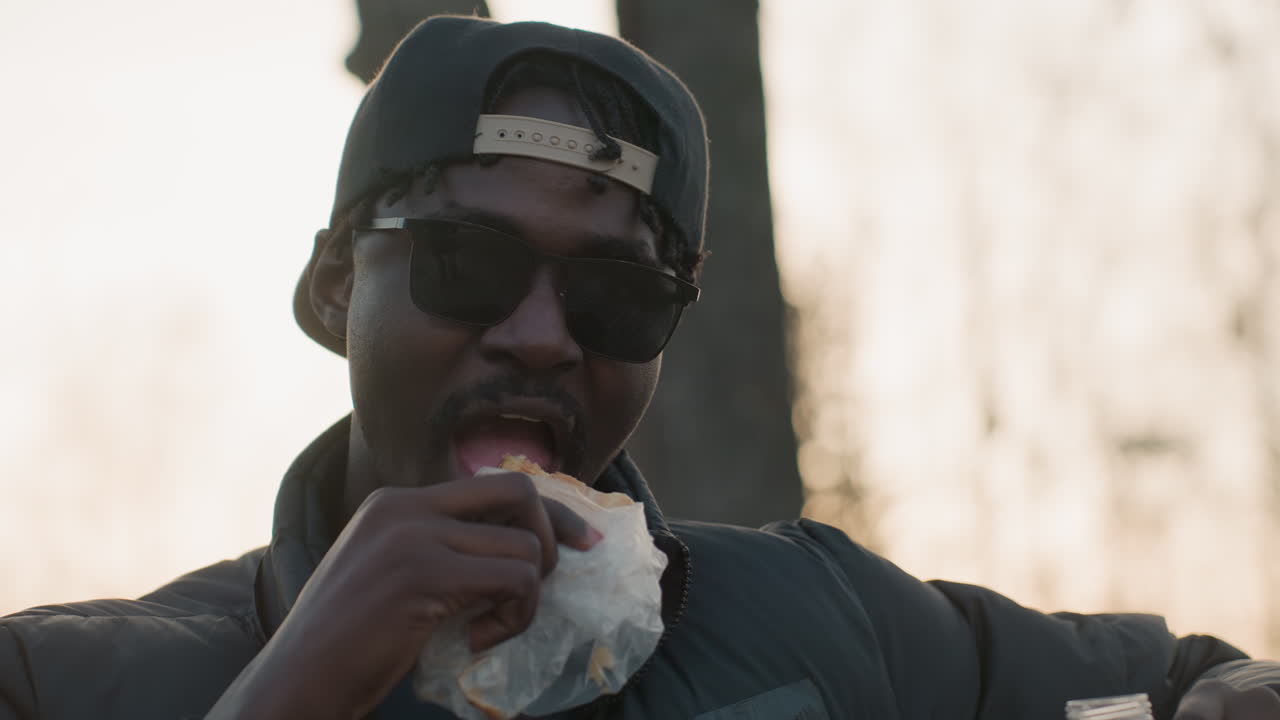 Close up of young man wearing sunglasses and backward cap enjoying bite of doughnut wrapped in paper outdoors during golden hour, casual jacket, relaxed expression, blurred background