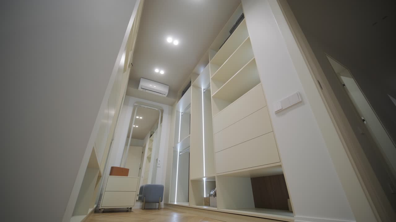 una sala de vestuario bien organizada muestra armarios altos y modernos con amplias estanterías e iluminación integrada, creando un área de almacenamiento de ropa sofisticada y funcional con una cómoda silla para vestirse.