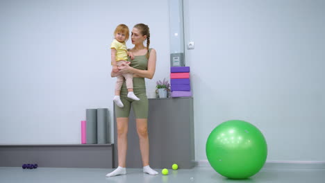 mother in workout gear holding toddler daughter in gym, with green exercise ball and purple dumbbells scattered around, attempting to gain child attention for playful bonding