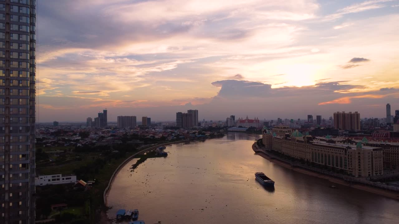 Drone descends to beautiful Tonle Bassac river, Koh Pich buildings under dusky sky with sunlight breaking through clouds in Phnom Penh, Cambodia.