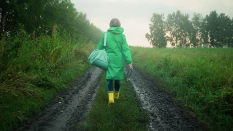 back view of woman in green raincoat carrying mint-colored bag and umbrella, wearing yellow rain boots, walking along a muddy path surrounded by tall greenery, trees, and a misty sky