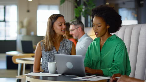 two businesswomen working on laptop in informal seating area of modern office on coffee break