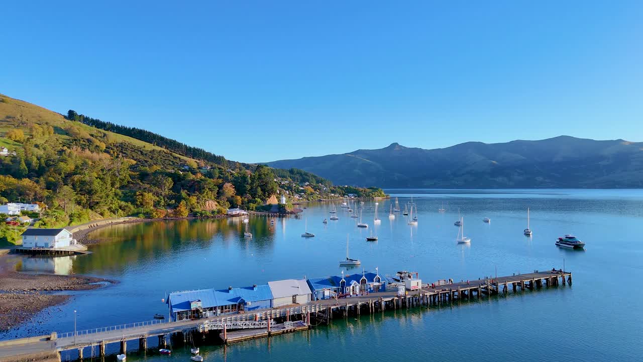 Aerial view of Akaroa Harbour with calm waters, boats, and surrounding hills under clear blue skies