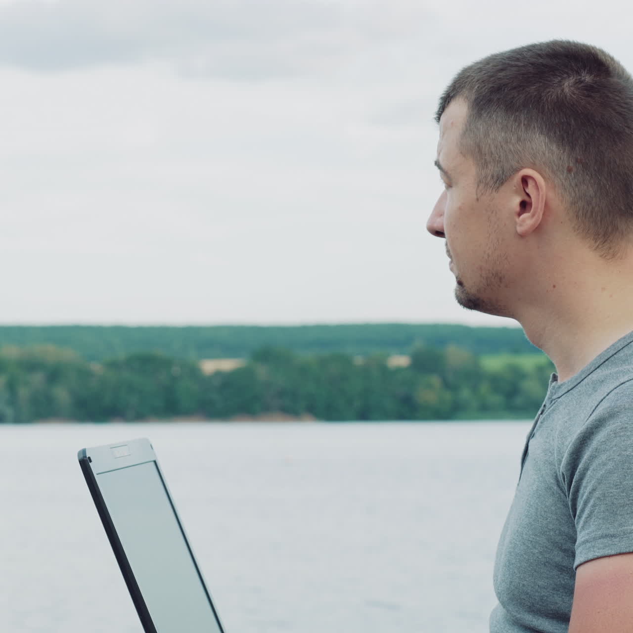 Handsome man typing on a laptop sitting over the flowing river and enjoys the nature in summer. Young male combines his work with a rest outdoors. Close-up