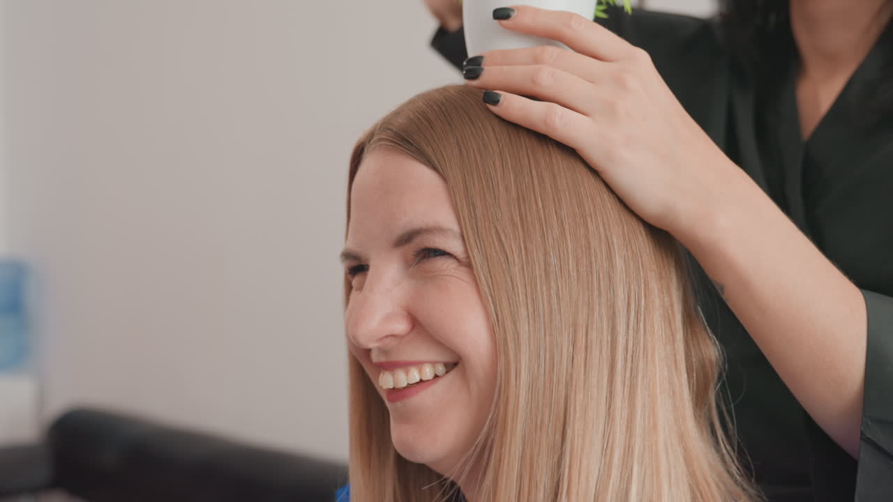 Una mujer caucásica sonríe en un salón de belleza mientras una estilista le coloca suavemente una planta en maceta sobre la cabeza, ambas riendo, luz de estudio acogedora, primer plano del cabello rubio, manos con manicura negra visibles, actitud lúdica y natural