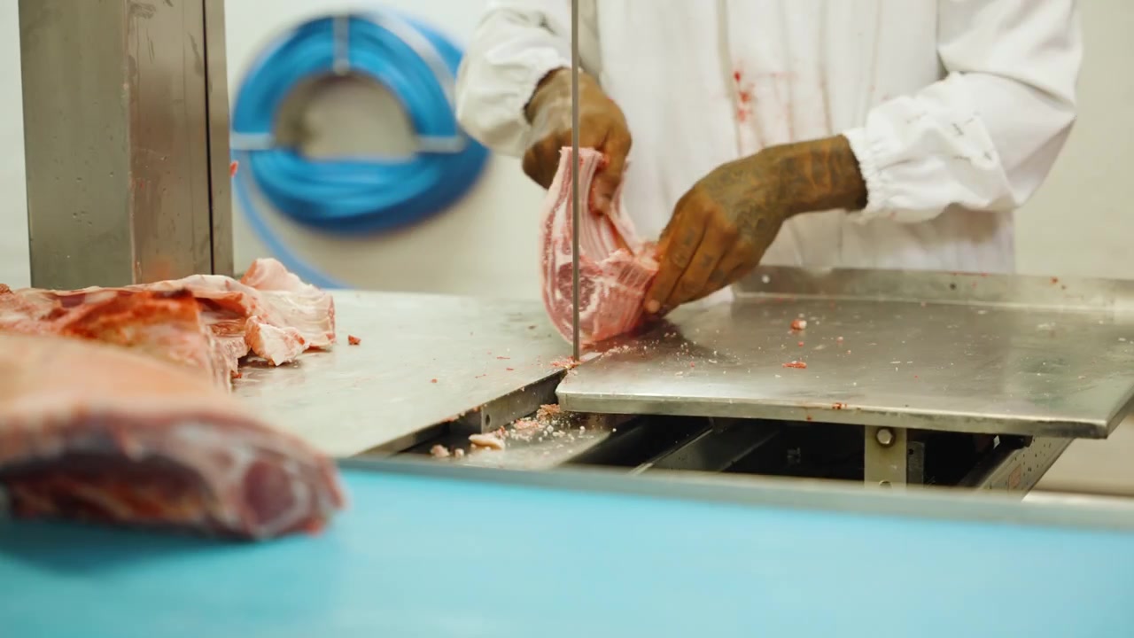 Meat processing worker cuts a pork carcass into ribs and sections on a stainless steel bandsaw table in an industrial butchery.