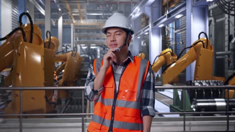 asian male engineer with safety helmet thinking about something and looking around while standing with industrial robotic arms working