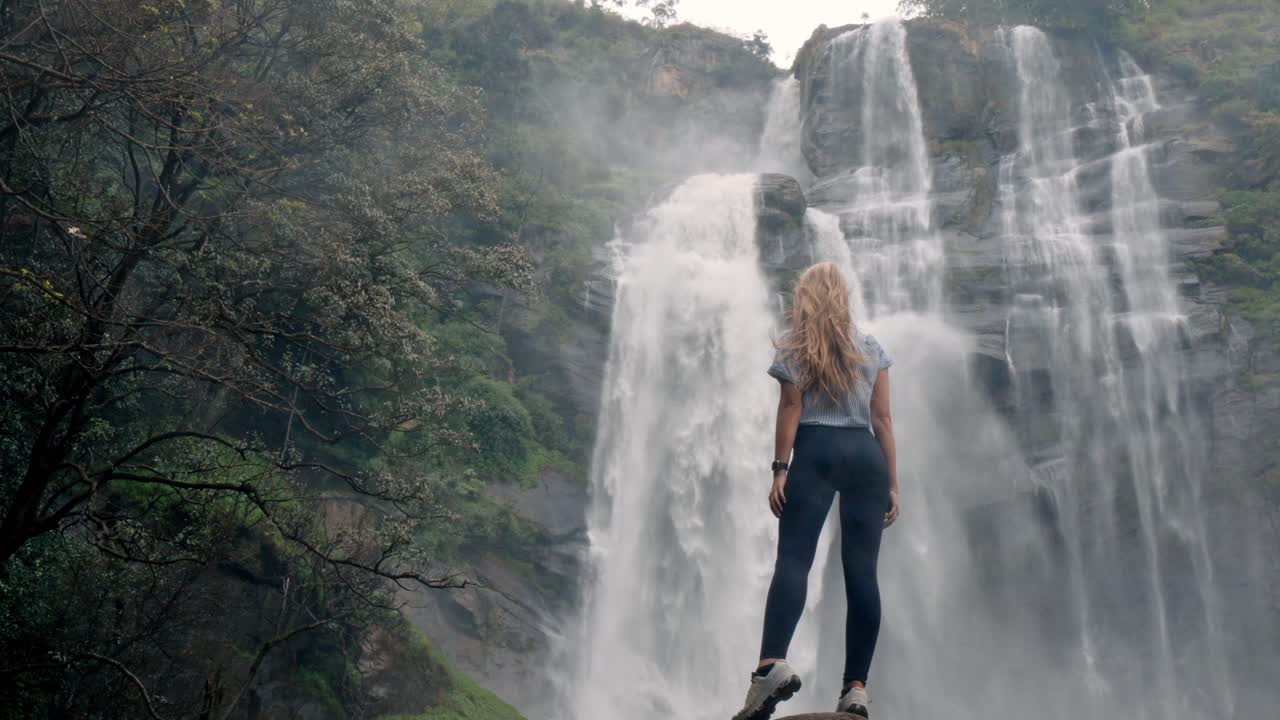 A woman stands in awe before the roaring Bomburu Ella Waterfalls, surrounded by mist, jungle, and rocky cliffs in the lush highlands of Nuwara Eliya, Sri Lanka.