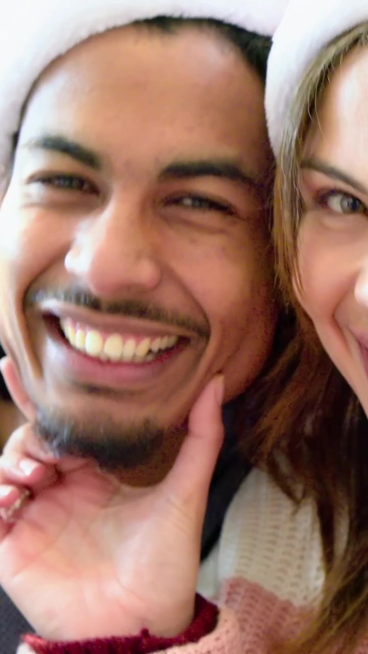 Video vertical: Cámara selfie grabando a una pareja ajustándose gorros de Papá Noel, riendo y apoyando la barbilla en la mano, con suéter en casa.